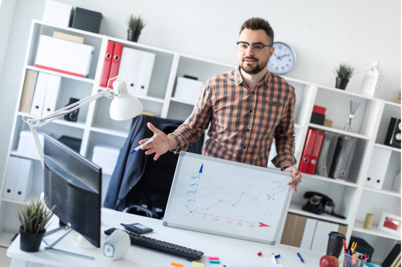 The man in the office is standing near the table holding a white board